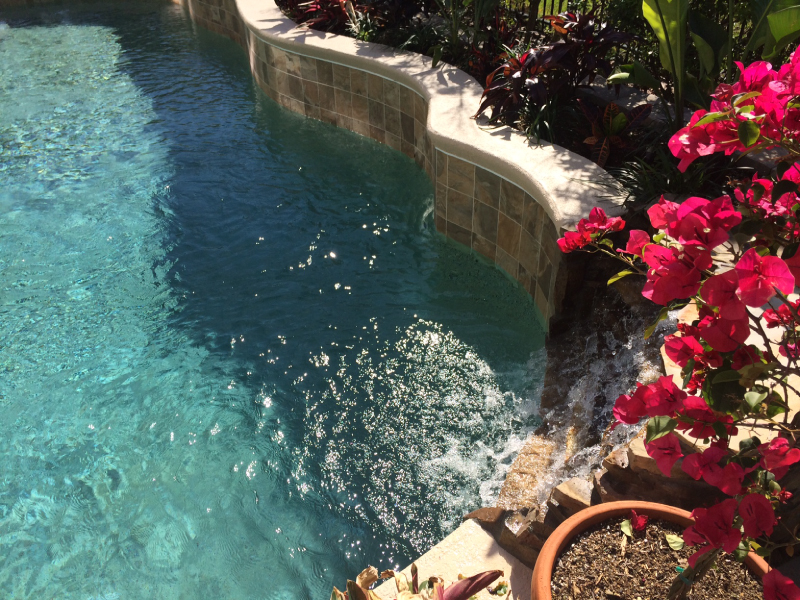 Close-up of a tiled raised wall with water spilling into a pool, framed by bright pink bougainvillea flowers.
