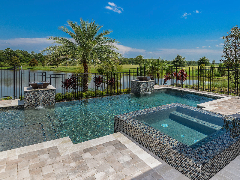 A tiled infinity-edge style pool with a raised spa and two water bowl fountains overlooking a lake and golf course.