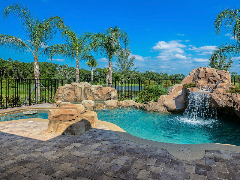 Freeform pool with a rock waterfall, slide, and grotto surrounded by palm trees under a black metal fence line.