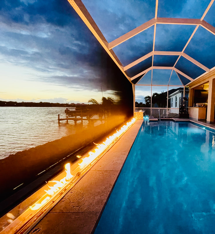 A long linear fire feature burns along the edge of a screened pool overlooking a waterway at dusk.