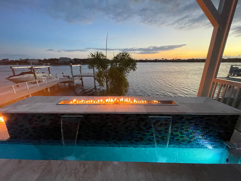 A linear fire feature burns atop a tiled wall with two water scuppers spilling into a lit pool at dusk on the waterfront.