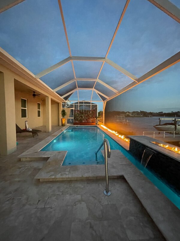 An L-shaped pool with a linear fire trough and waterfall spillway overlooking a canal at dusk under a screened enclosure.