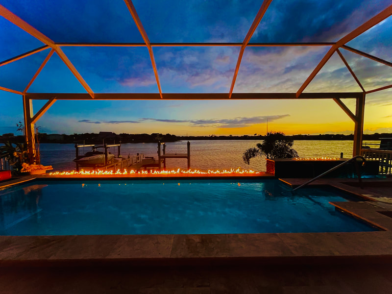 A screened waterfront pool with a linear fire feature and raised spa glowing at dusk over the water.