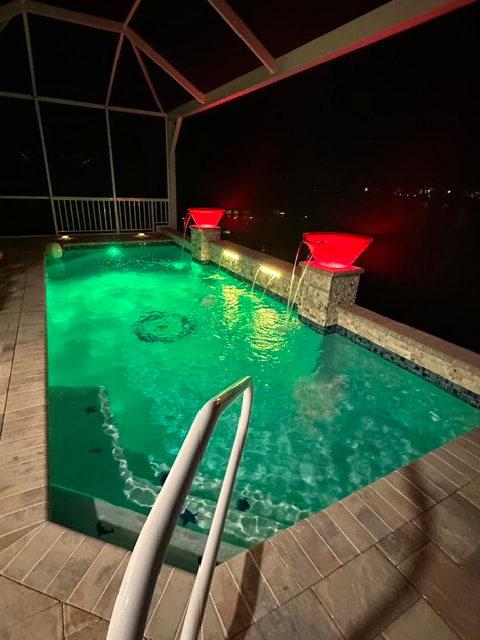 A screened backyard pool glowing green at night with two red-lit water spouts overlooking a canal.