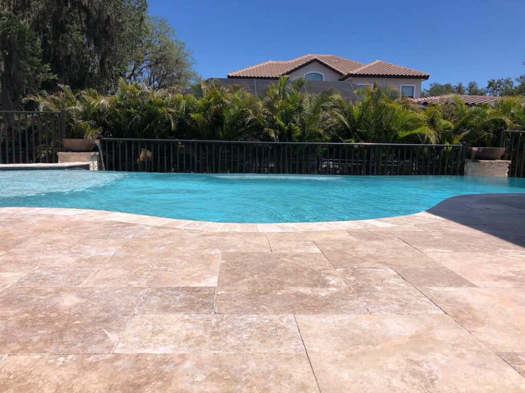 Freeform pool with travertine deck and two stone water bowls spilling into the water, backed by a metal fence and palm trees.