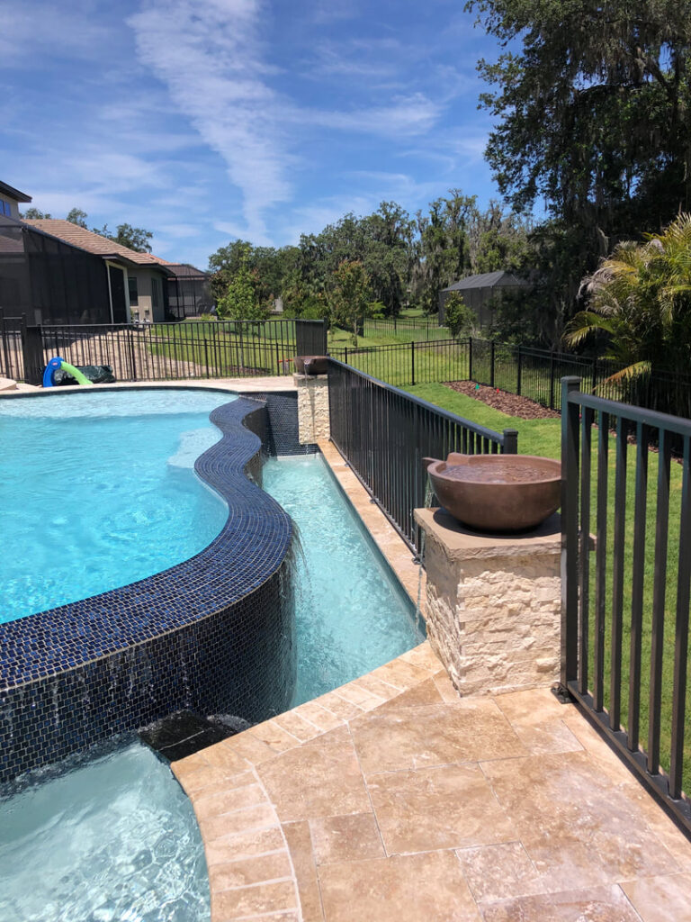 Freeform pool with raised spa spilling over a dark mosaic tile wall onto a travertine deck.