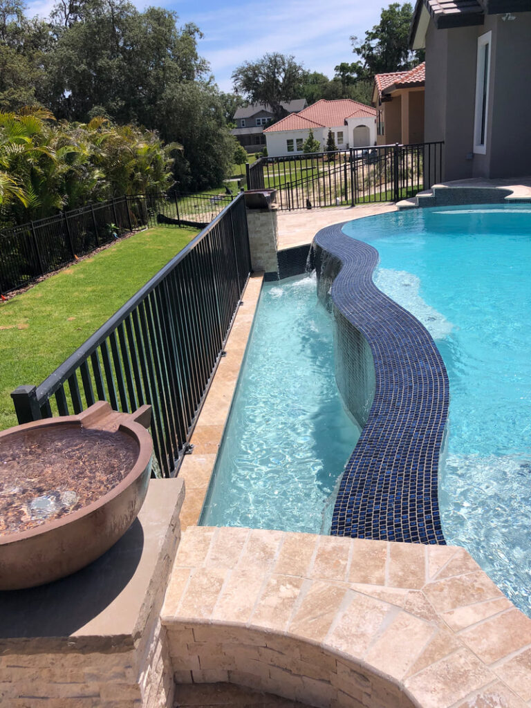 A tiled water channel with a spillway feature runs beside a curving pool edge, with a decorative water bowl in the foreground.
