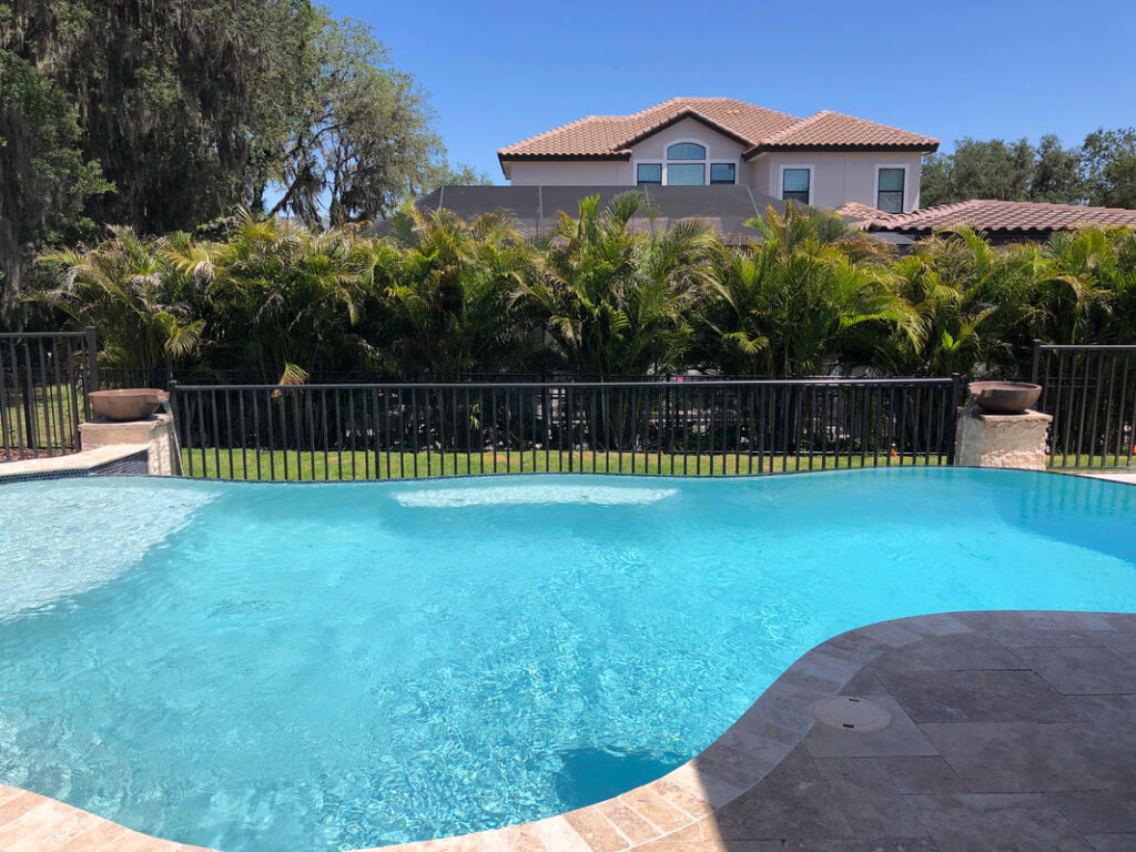 A freeform pool with travertine decking, seen partially with a tall home and palm trees behind a black metal fence.