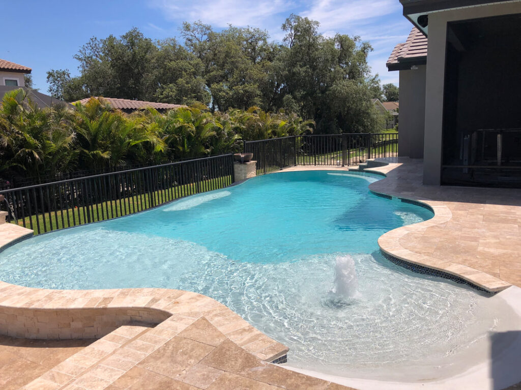 A freeform swimming pool with a bubbler fountain and travertine deck, enclosed by a black metal fence with palm trees in the background.