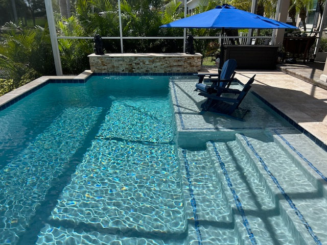 A small rectangular pool with a broad tanning ledge holding two submerged chairs under a blue umbrella, enclosed in a screened lanai.