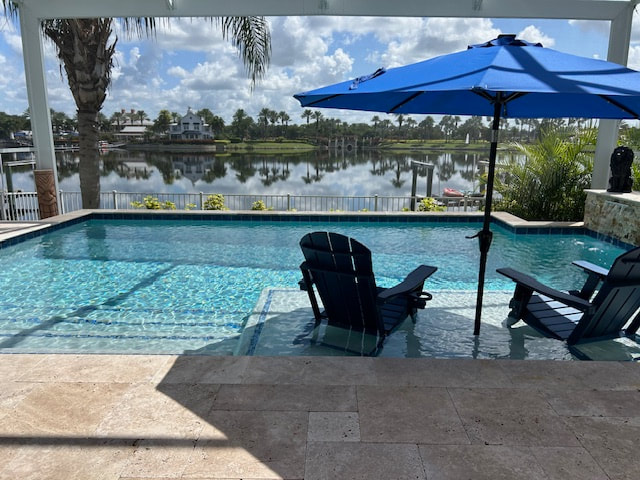A small freeform pool with a shallow tanning ledge and loungers overlooking a calm lakefront under a blue patio umbrella.