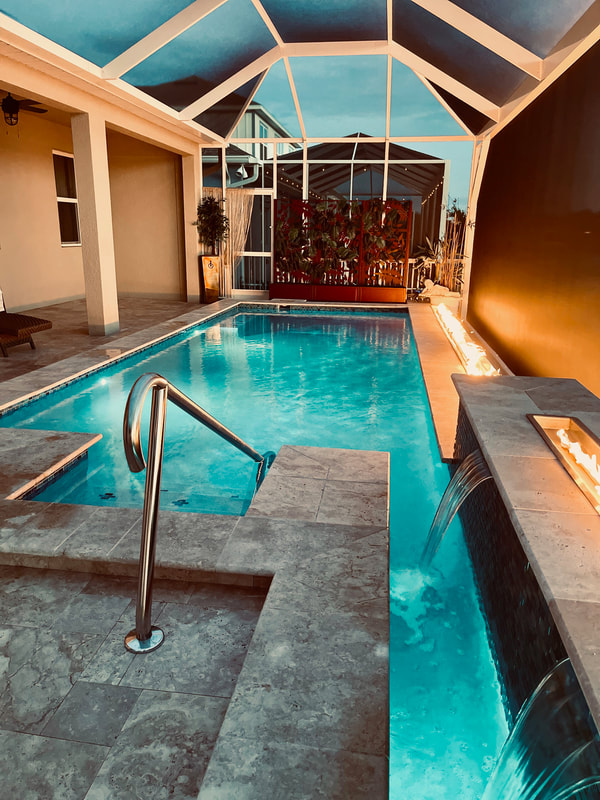 A rectangular backyard pool with a raised spa and a linear fire trough spilling water into the pool at dusk under a screened enclosure.