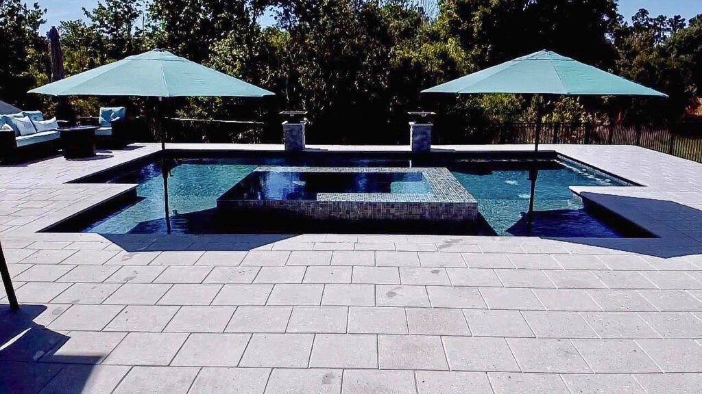 A rectangular backyard pool with a raised tiled spa in the center, surrounded by a paver patio and shaded by two umbrellas.