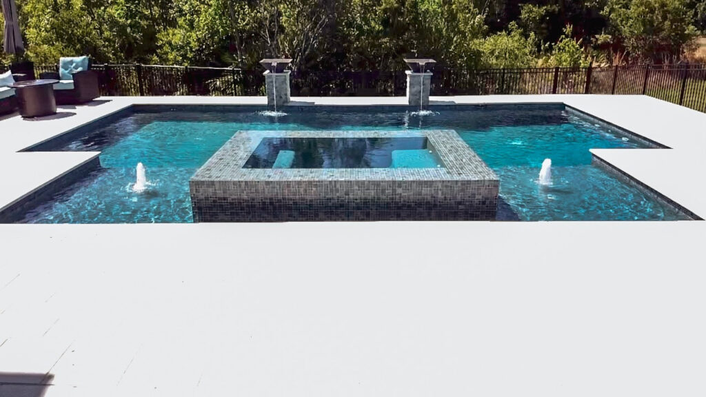 A rectangular backyard pool with a raised tiled spa in the center and two deck-jet fountains spraying into the water.