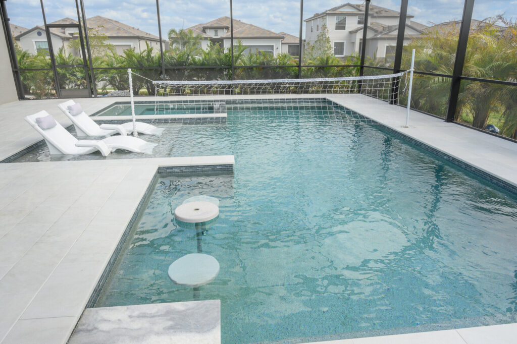 An L-shaped screened pool with in-water loungers, a volleyball net, and floating table stools.