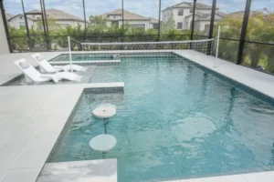 An L-shaped screened pool with in-water loungers, a volleyball net, and floating table stools.