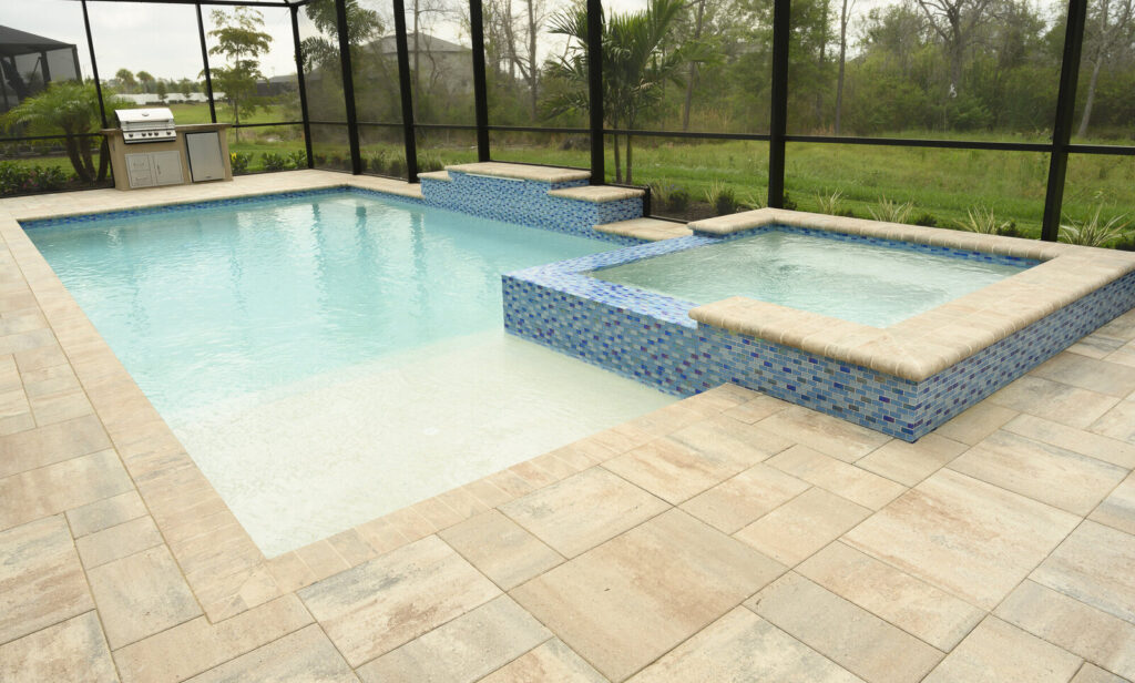 A rectangular pool with a raised tiled spa and stone-paved deck under a screened enclosure.