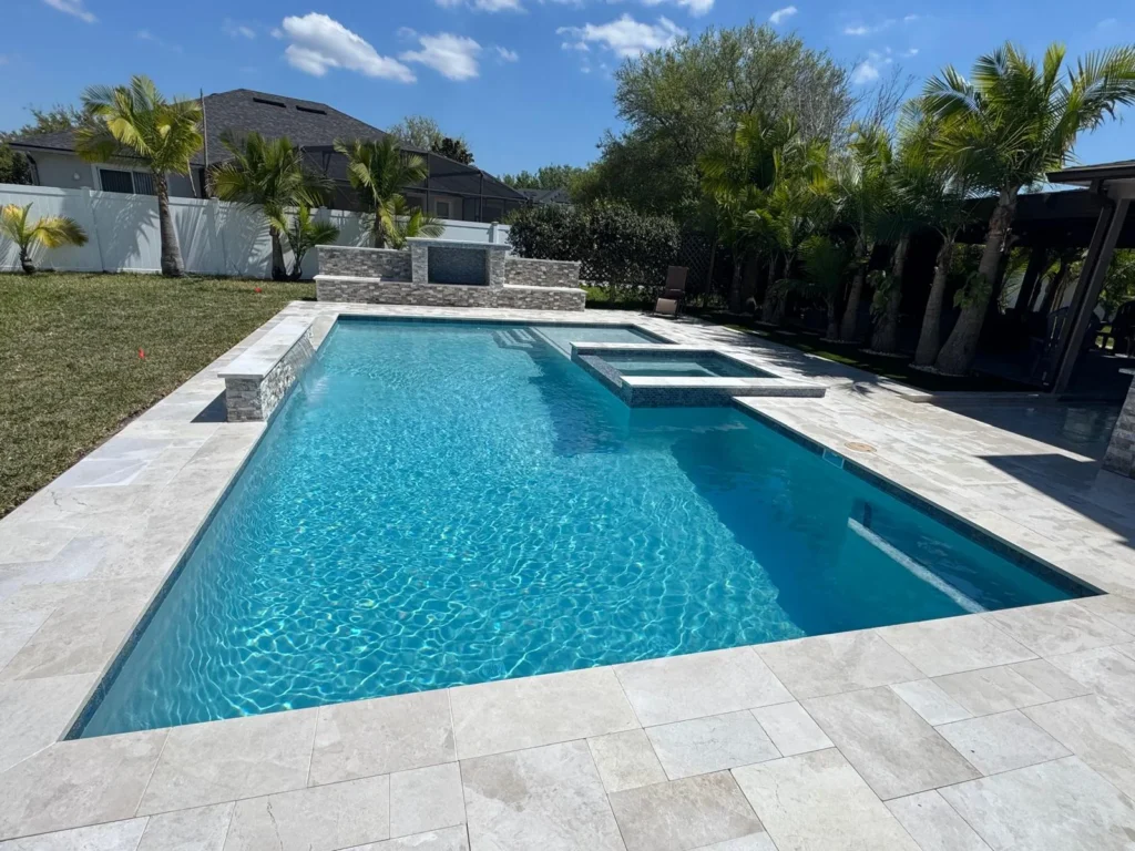 A rectangular gunite pool with an attached raised spa and stone water feature wall, surrounded by travertine pavers and palm trees.