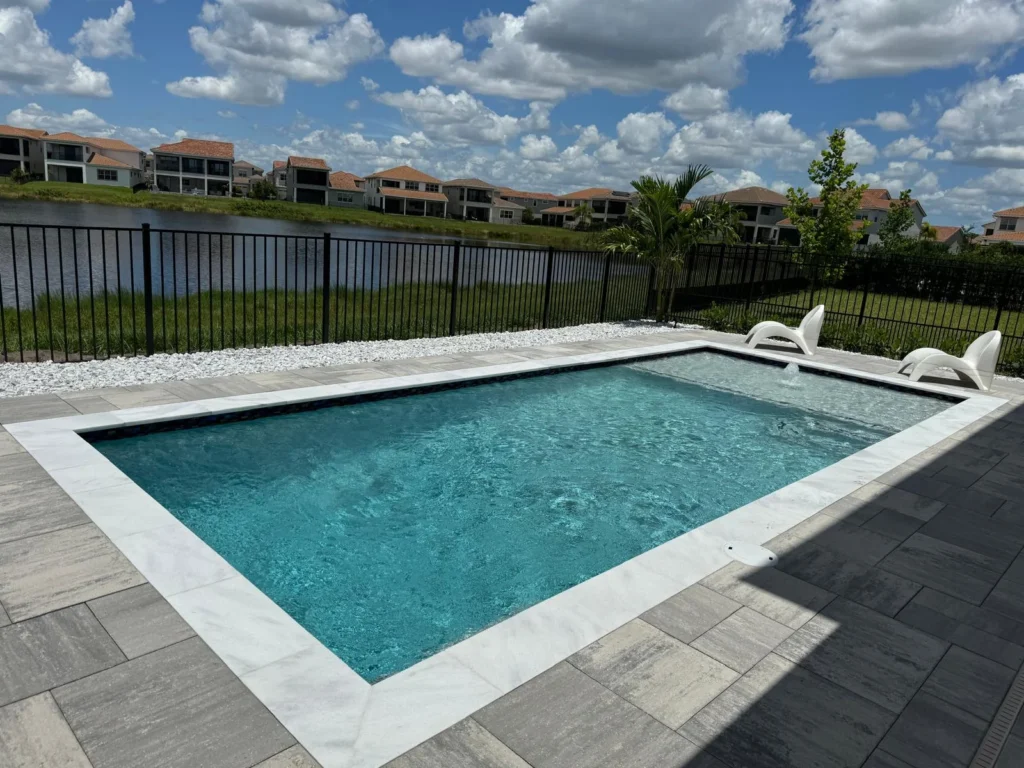A rectangular backyard pool with light stone coping and paver deck, overlooking a lake with houses in the distance.