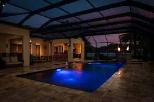 A screened lanai pool glowing blue at dusk with deck jets and twin fire bowls beside a stone waterfall.
