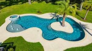 Aerial view of a freeform pool with a raised spa, fountain jets, and a central island with a palm tree, surrounded by a travertine deck and lawn.