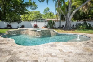 Freeform backyard pool with a raised stone spa and spillover waterfall on a travertine paver patio.