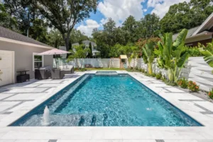 A rectangular backyard pool with a raised spa and fountain jets, surrounded by white marble-look pavers and lush tropical landscaping.