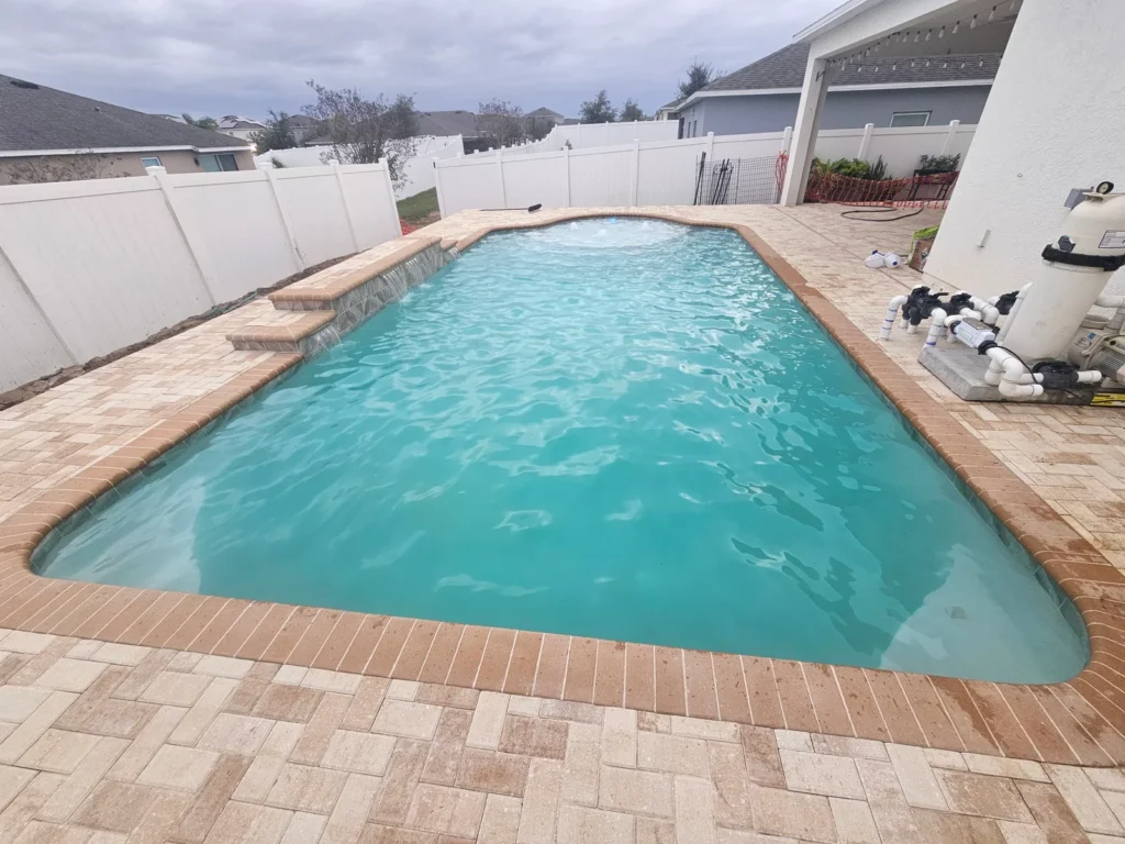 A rectangular backyard pool with a small raised water feature, surrounded by travertine pavers and a white vinyl fence under an overcast sky.