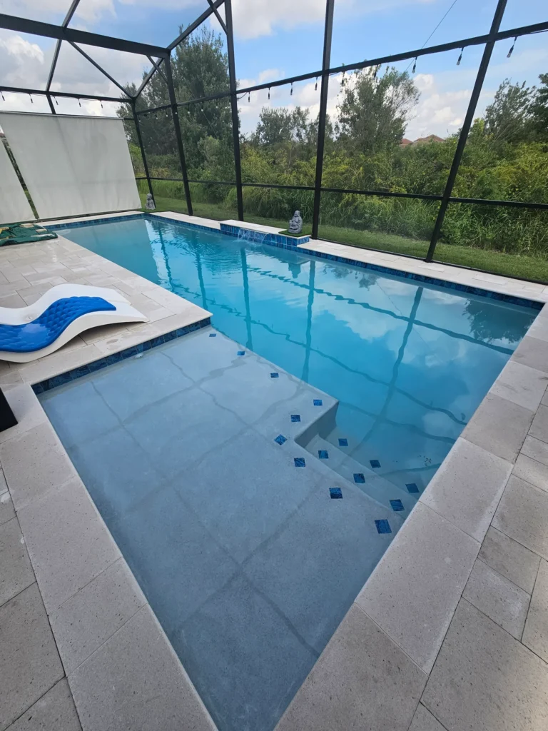 A rectangular screened pool with an attached shallow sun shelf, travertine deck, and small statue water feature.