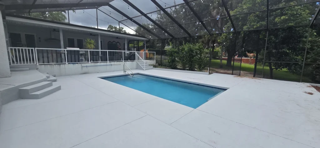 A small rectangular pool with a large white concrete deck under a screened lanai.
