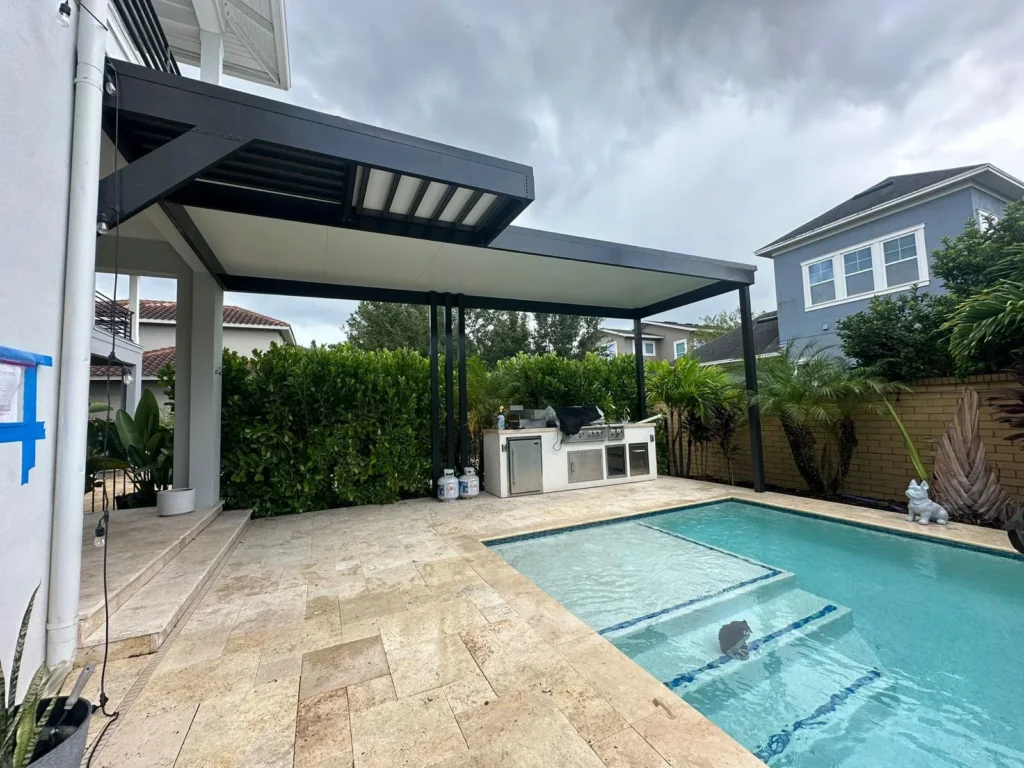 A corner of a backyard pool with entry steps, seen beside a travertine patio and outdoor kitchen under a modern pergola.