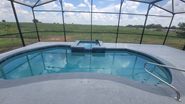 A kidney shaped vinyl pool with a raised spa under a screen enclosure, overlooking open fields.
