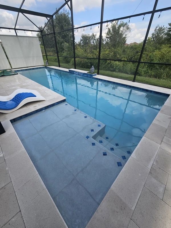 A rectangular pool with a shallow tanning ledge under a screened lanai, framed by travertine pavers.