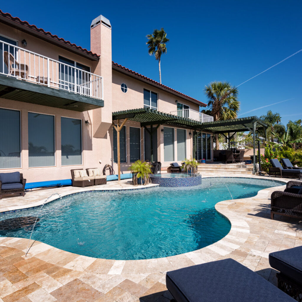 Freeform backyard pool with raised tiled spa, water jets, and travertine deck beside a two-story stucco home.