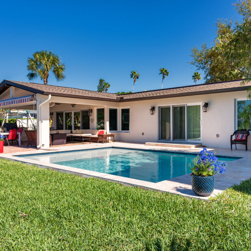 A small rectangular backyard pool with travertine deck in front of a ranch-style home under a clear blue sky.