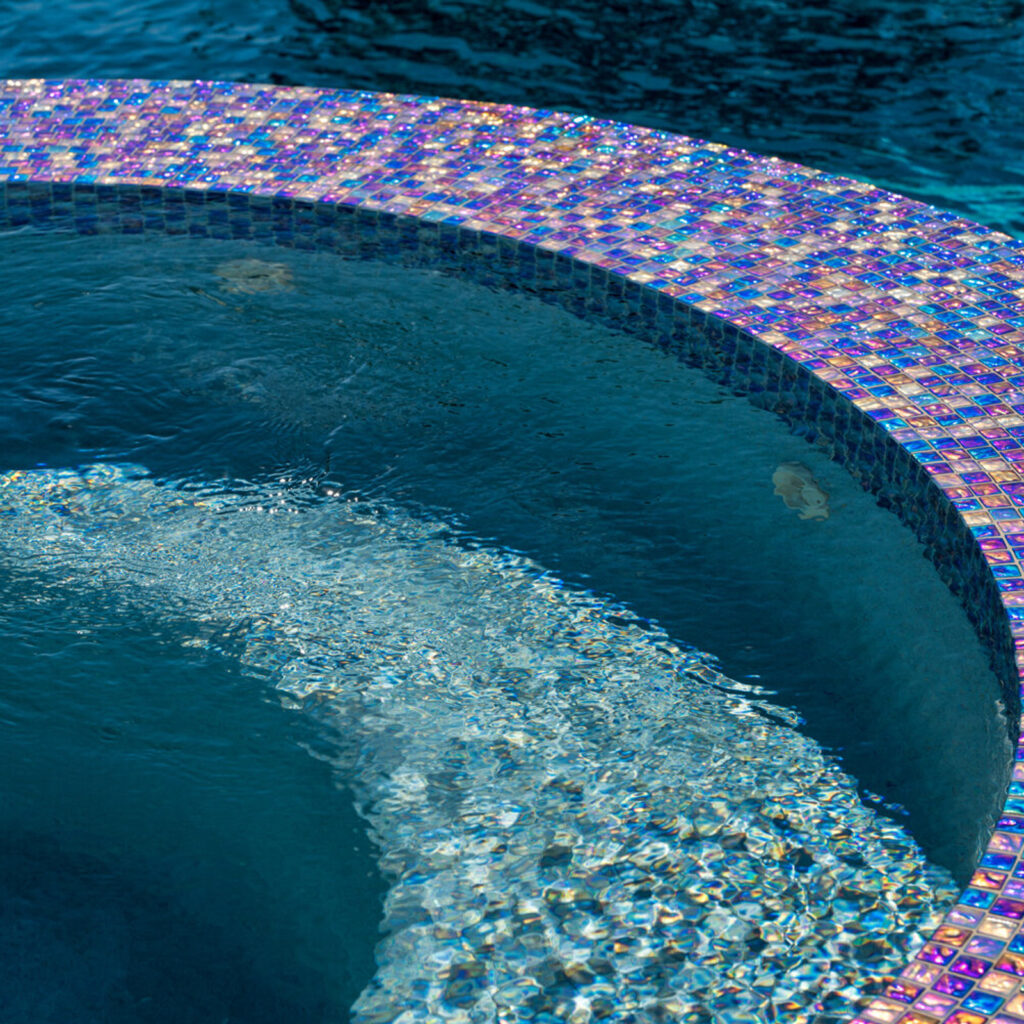Close-up of a curved pool edge with iridescent glass mosaic tile and water spilling over a submerged step.