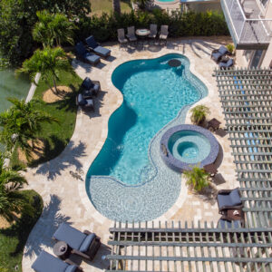 Aerial view of a freeform pool with a beach entry and a raised spa on a travertine patio beside a canal.