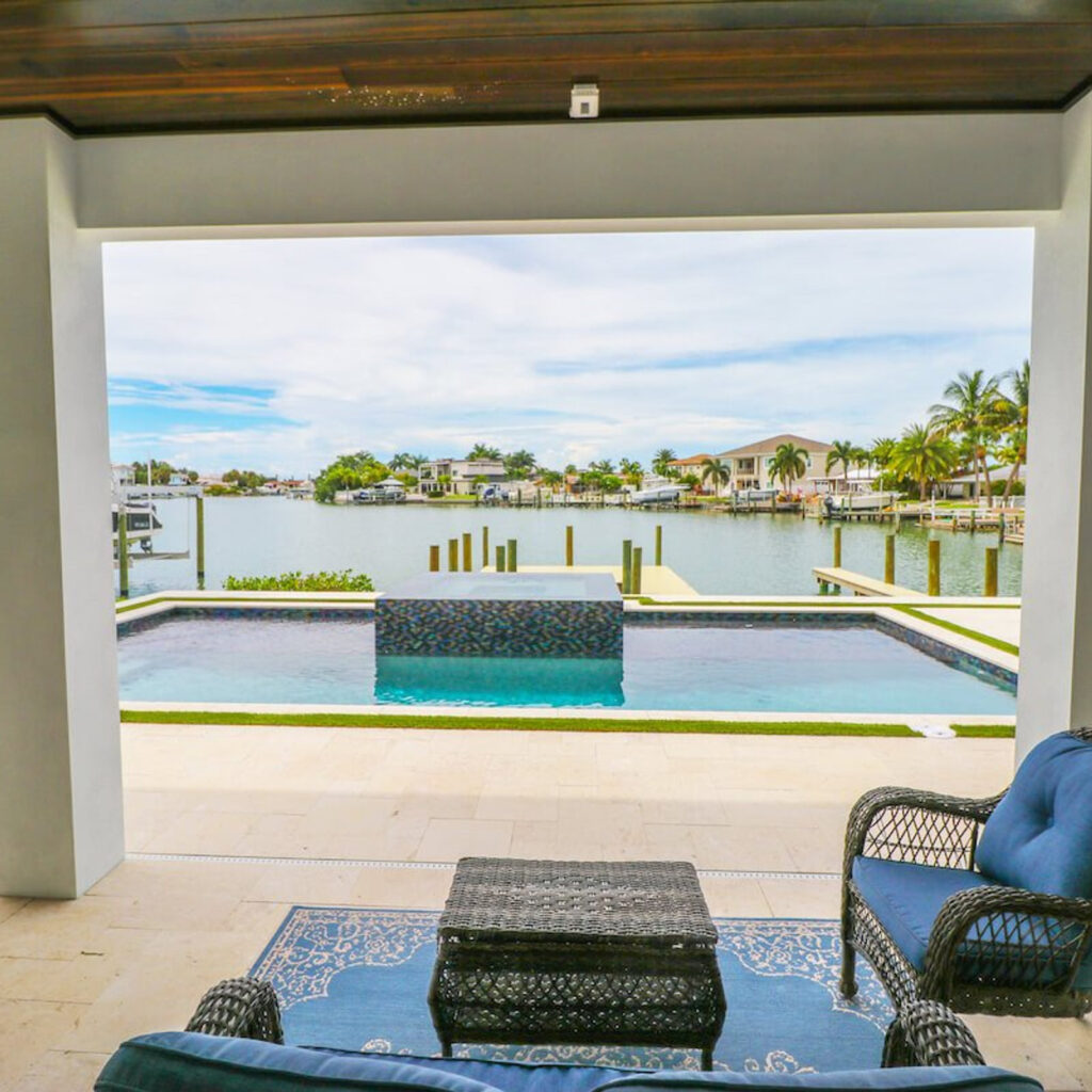 View from a covered lanai across an infinity-edge style pool with a raised tiled water feature toward a canal lined with docks and homes.