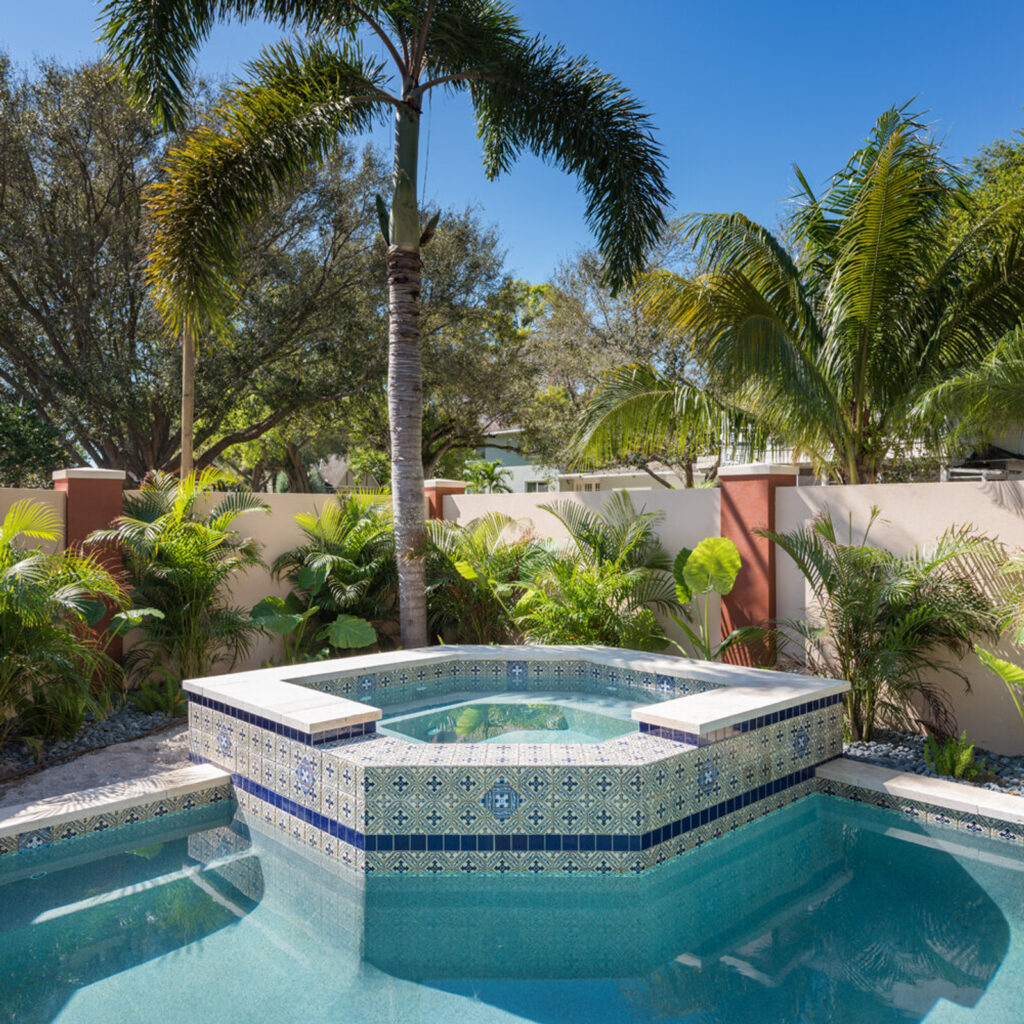 A raised octagonal spa with ornate blue and white tile work sits beside a pool surrounded by palm trees and a privacy wall.
