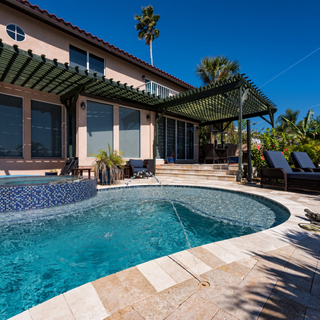 A freeform pool with a raised tiled spa spilling water over the edge, surrounded by a travertine paver patio under a pergola.