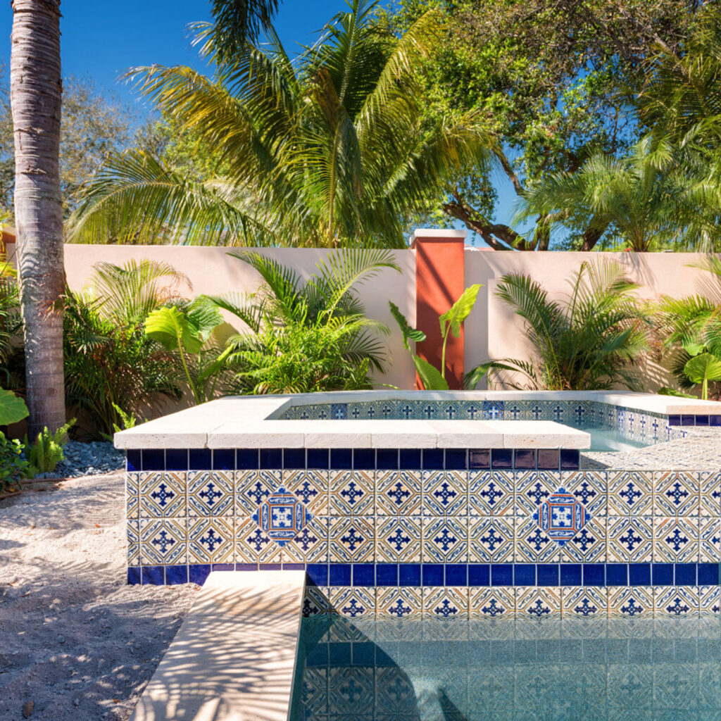 A raised spa wall covered in ornate blue and white Talavera-style tile beside a pool edge in a tropical backyard.