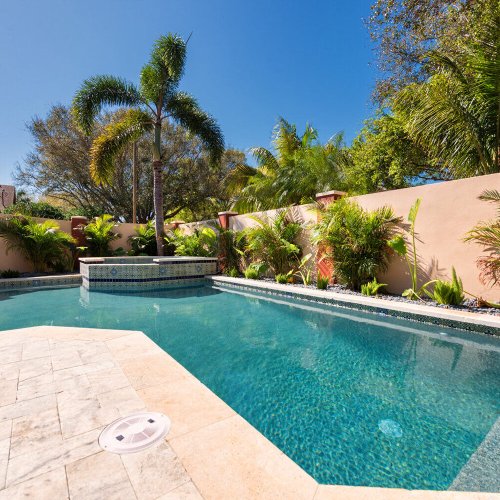 A rectangular backyard pool with a raised tiled spa in the corner, surrounded by a travertine patio and tropical landscaping against a stucco wall.