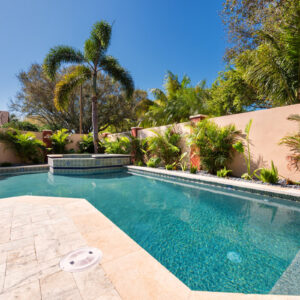 A rectangular backyard pool with a raised tiled spa in the corner, surrounded by a travertine patio and tropical landscaping against a stucco wall.