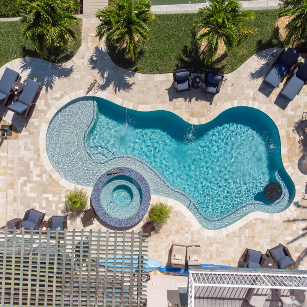 Aerial view of a freeform pool with a tanning ledge and round attached spa surrounded by travertine decking and palm trees.