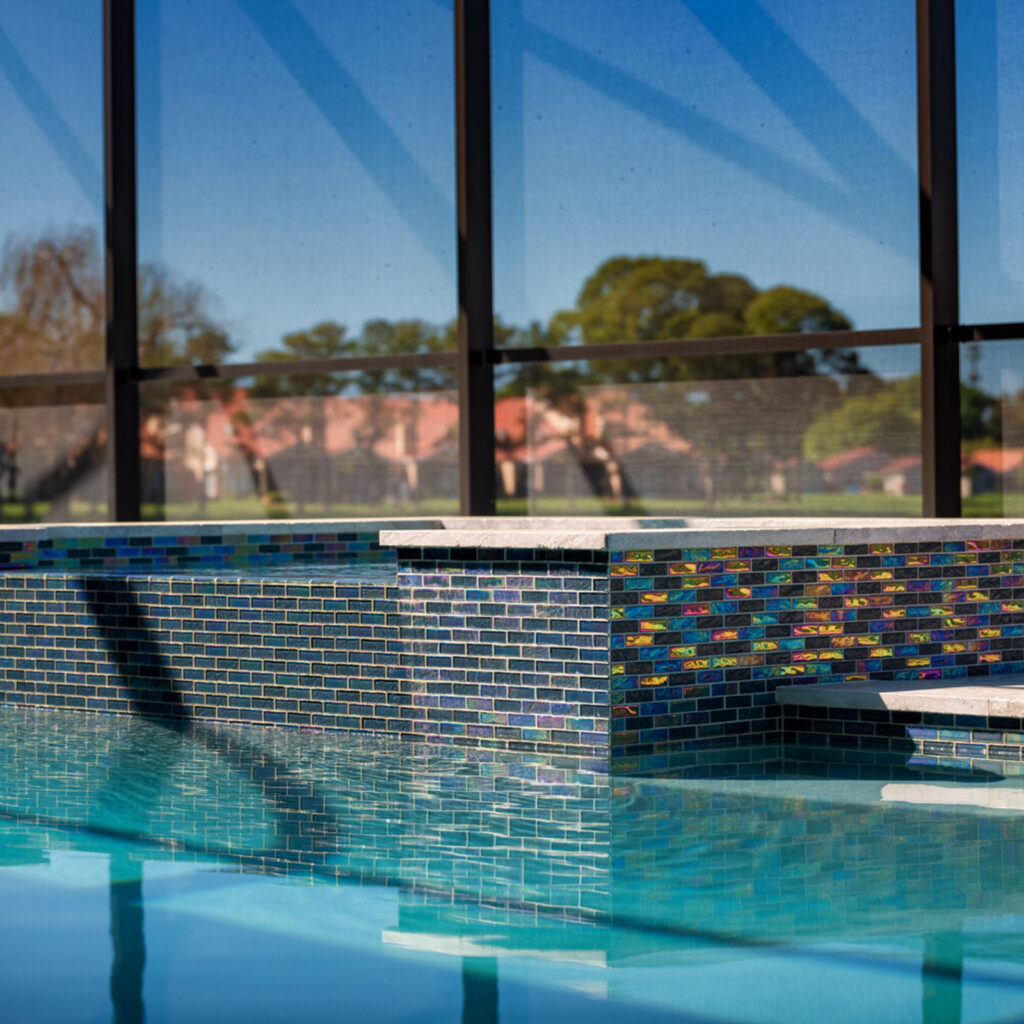 A pool corner with a raised wall covered in iridescent glass mosaic tile under a screened enclosure.
