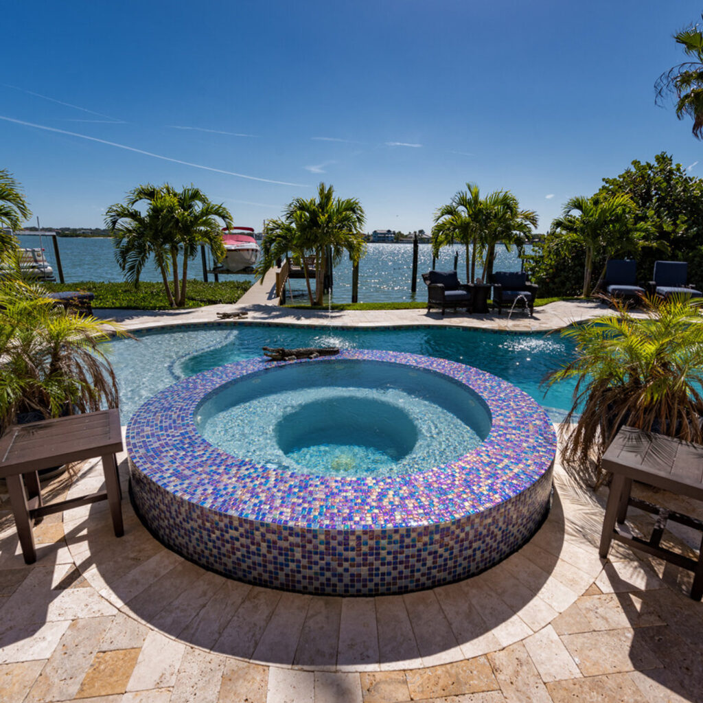 A raised round spa covered in iridescent mosaic tile sits in front of a freeform pool overlooking a waterway with docked boats.
