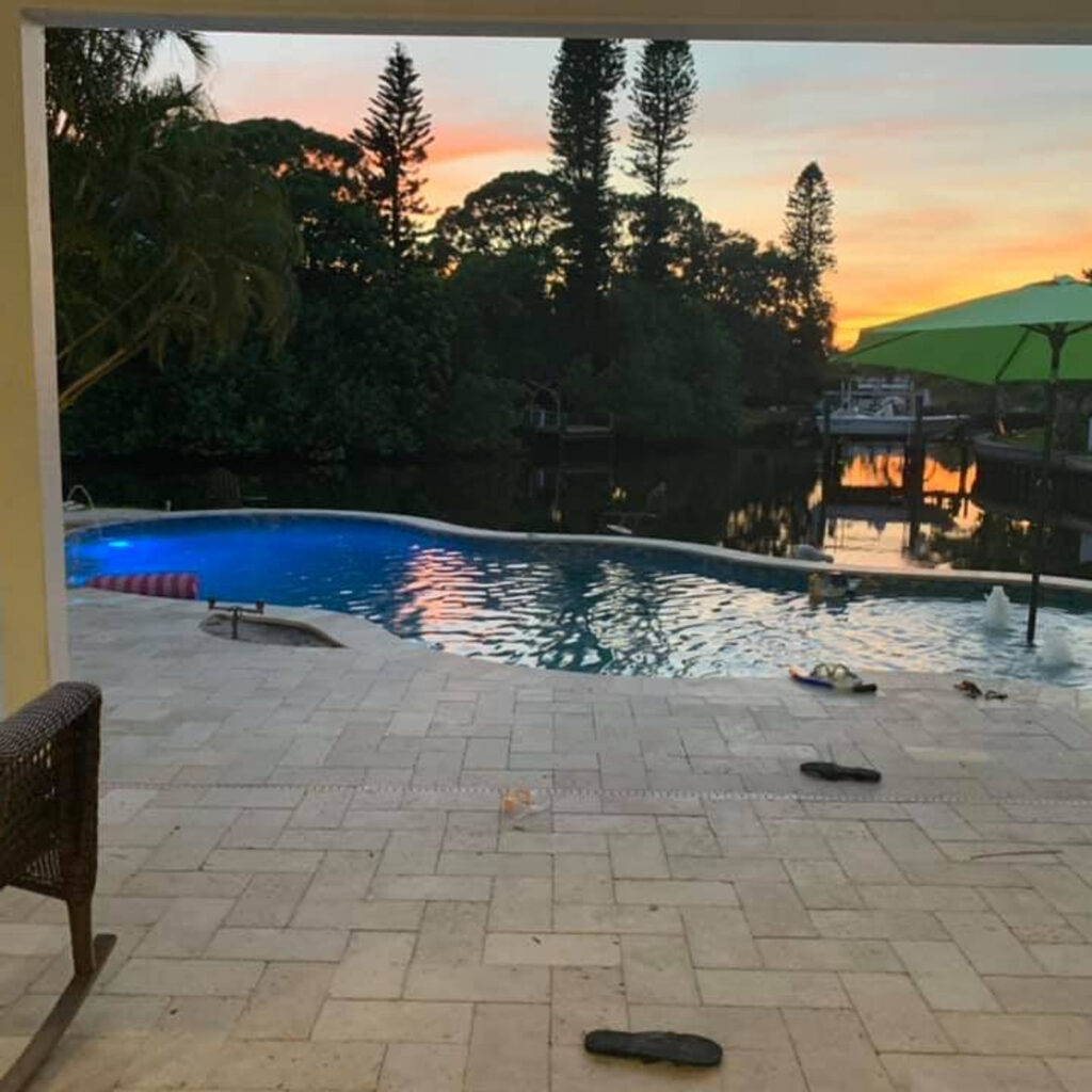 Freeform pool on a canal at sunset with a travertine paver patio and distant figures swimming.