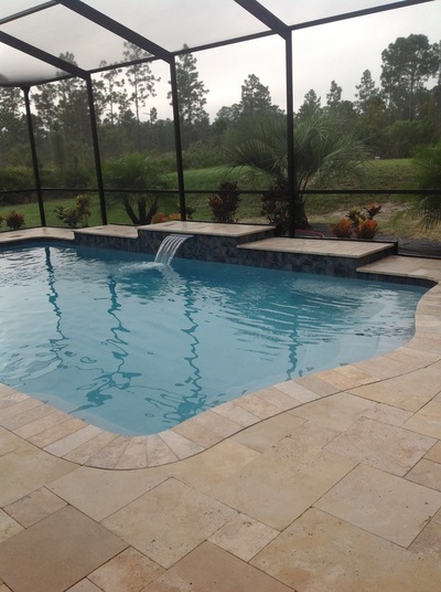 A freeform backyard pool with a raised tiled wall water feature under a screened lanai on an overcast day.