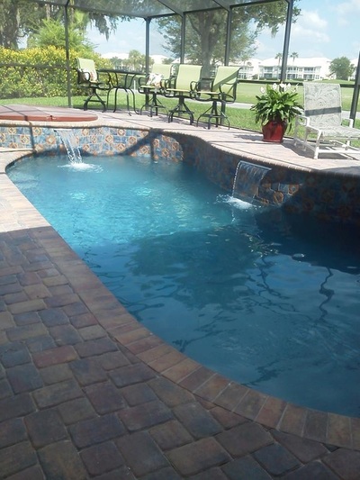 A freeform pool with a raised spa spilling water over tiled waterfalls onto a brick paver patio under a screen enclosure.