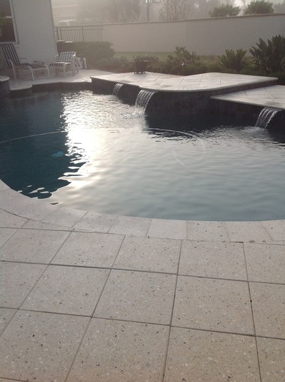 A freeform backyard pool with a raised wall featuring two water spillways, set on a stone paver patio under overcast sky.